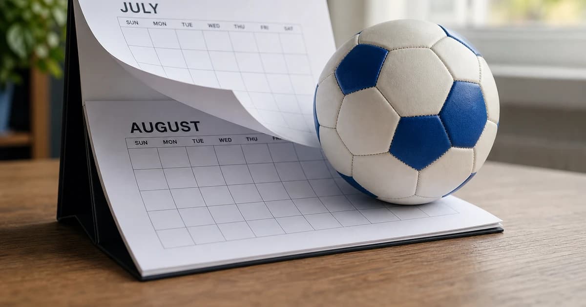 A wall calendar flipping from July to August with a soccer ball resting on the August page on a wooden desk illustrating the August 1 youth soccer age-group transition.